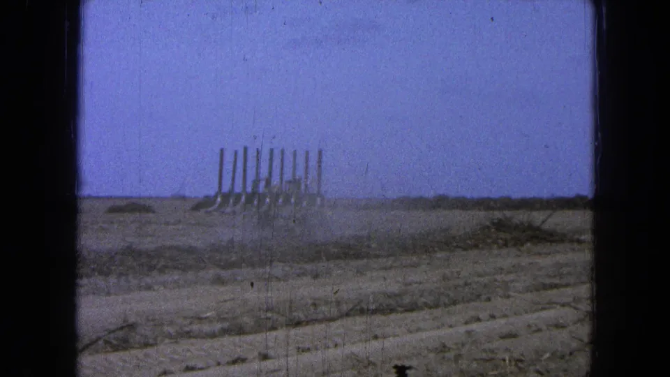 Distant 1965 Pueblo, Mexico view of a plow moving across a dry open field, with dust trailing near the ground and a low horizon beyond.