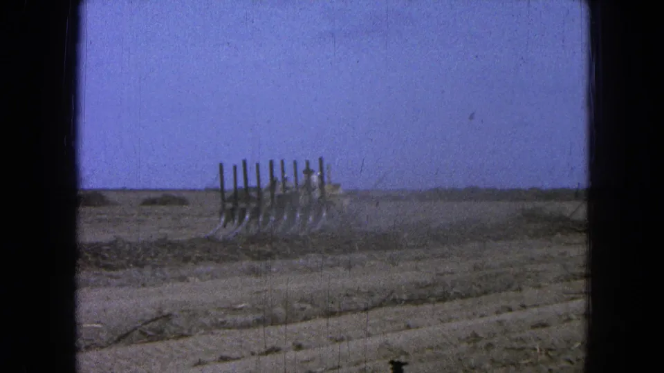 Final 1965 Pueblo, Mexico frame showing the plow in the field amid dust, furrowed soil, sparse brush, and a bright blue sky.