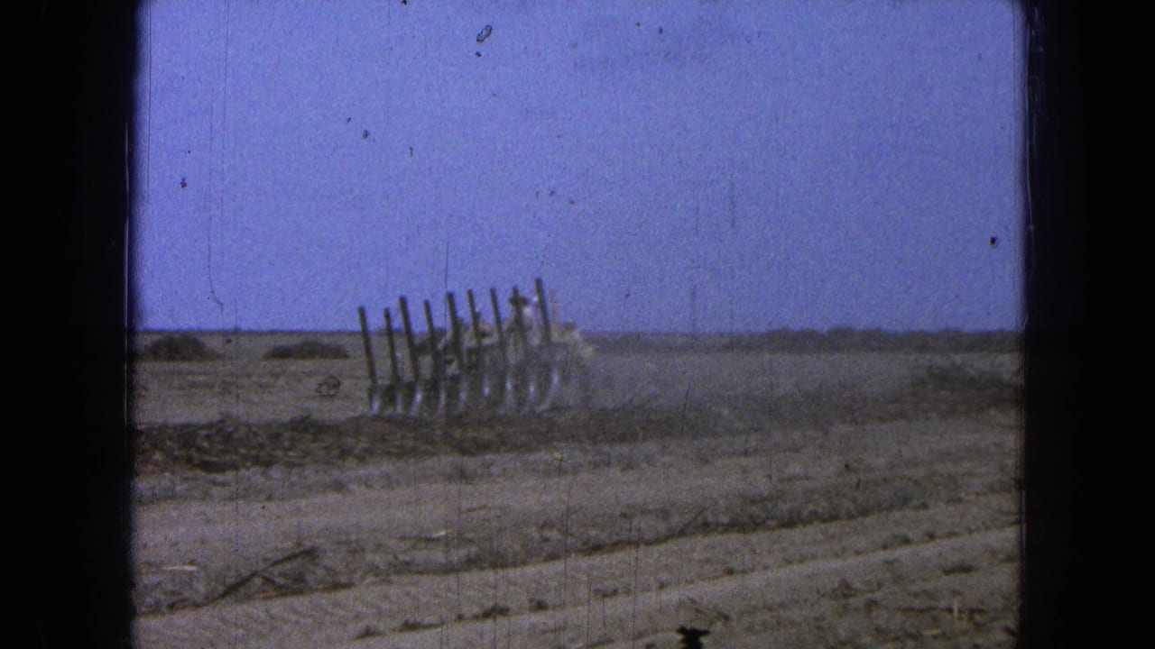 Distant tractor and fence posts in a dry open field under blue sky.