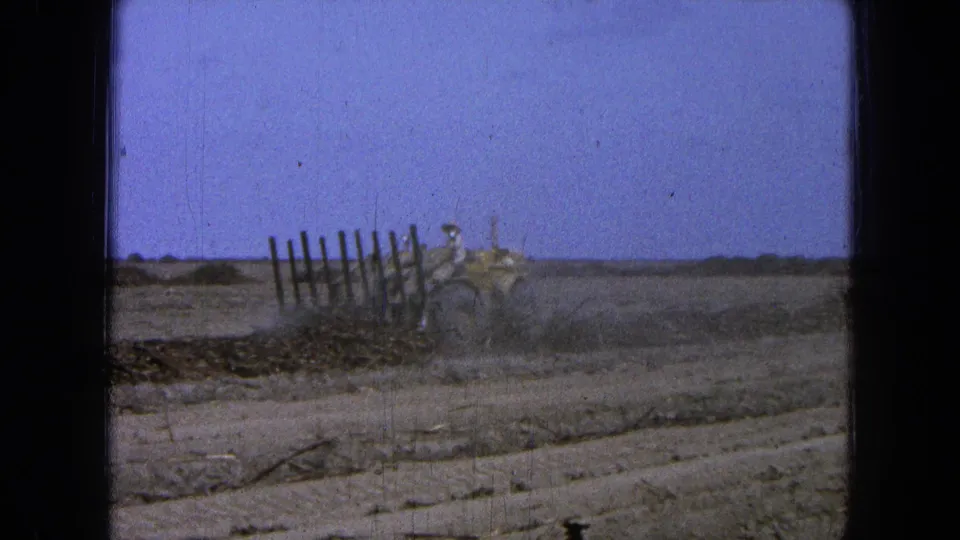 A distant operator drives a yellow tractor through dry soil, with churned dust and fence posts visible in the 8mm frame.
