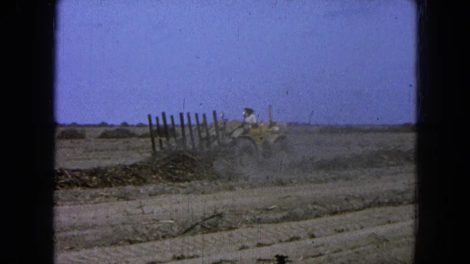 The tractor moves through the right side of the dry field, throwing up dust beneath a pale blue sky in the 1965 home movie.