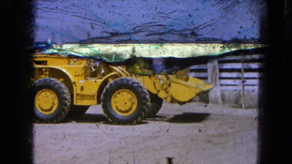 1965 Pueblo, Mexico 8mm frame showing a yellow front-end loader beside a wooden fence and simple structures on a dirt lot.