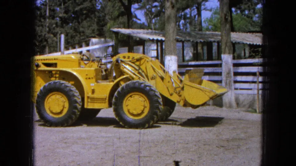 1965 Pueblo Mexico 8mm frame showing a yellow Caterpillar 922 loader in side profile on a dirt lot near a shed and trees.