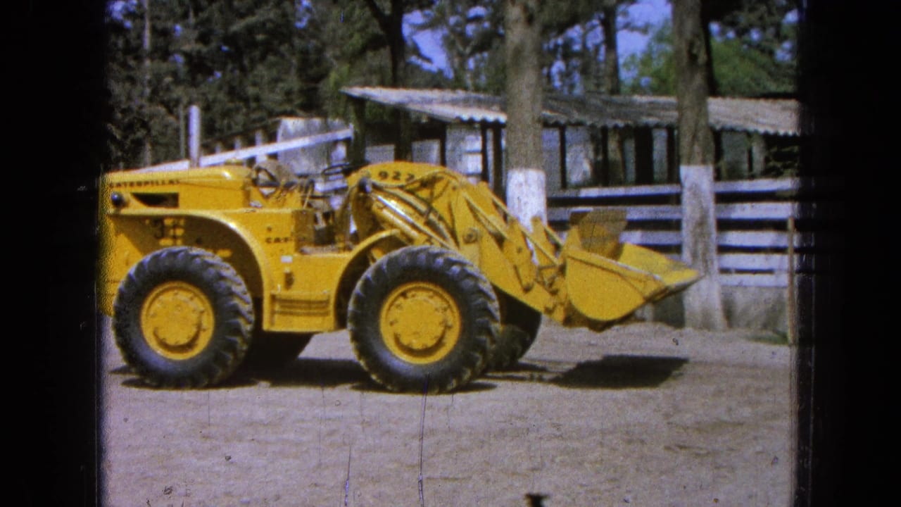 1965 Pueblo Mexico 8mm frame of a yellow Caterpillar loader marked 922 beside wooden rails, trees, and a low shed.