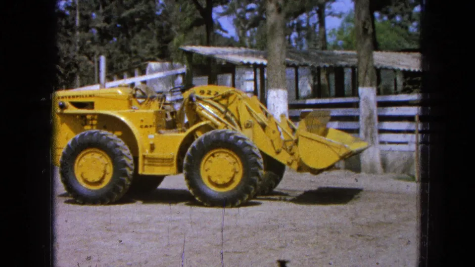 1965 Pueblo Mexico 8mm frame of a yellow Caterpillar loader marked 922 beside wooden rails, trees, and a low shed.