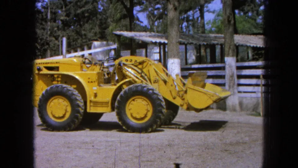 1965 Pueblo Mexico home-movie frame showing the parked Caterpillar loader from the side in a rural outdoor setting.