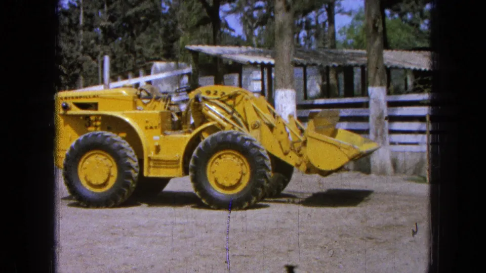 1965 Pueblo, Mexico footage shows a yellow Caterpillar front-end loader in profile near a white fence and corrugated-roof buildings.