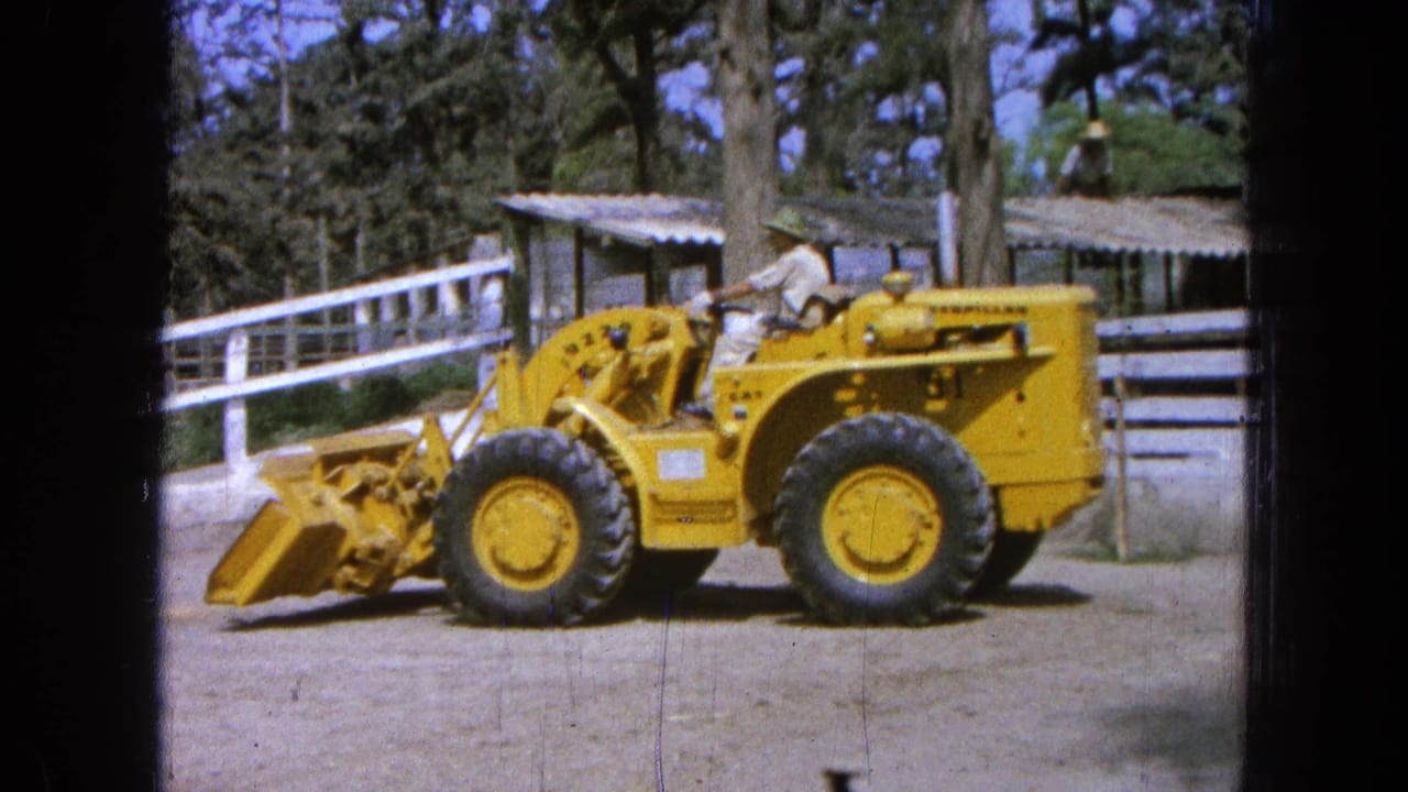 In Pueblo, Mexico, a worker in light clothing drives a yellow Caterpillar loader across a dirt yard with the bucket lowered.