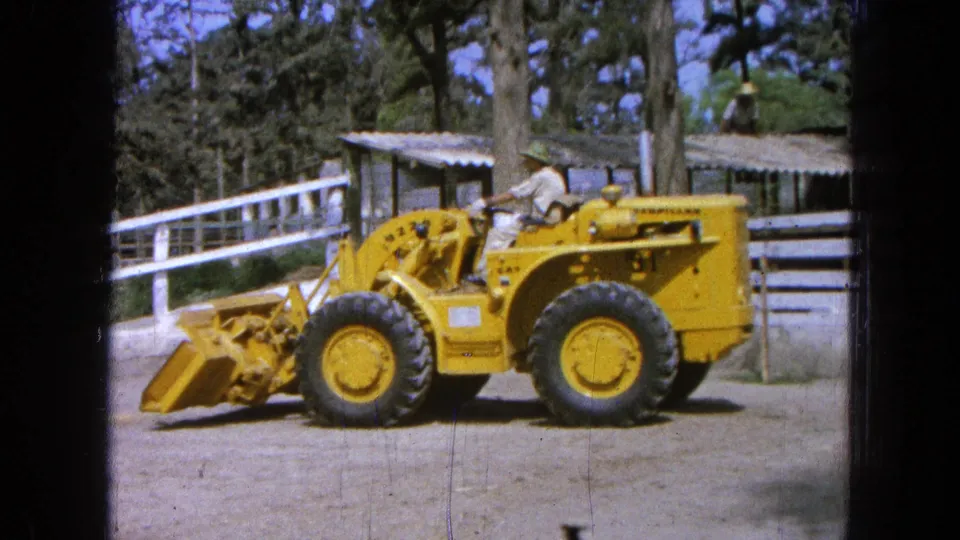 In Pueblo, Mexico, a worker in light clothing drives a yellow Caterpillar loader across a dirt yard with the bucket lowered.