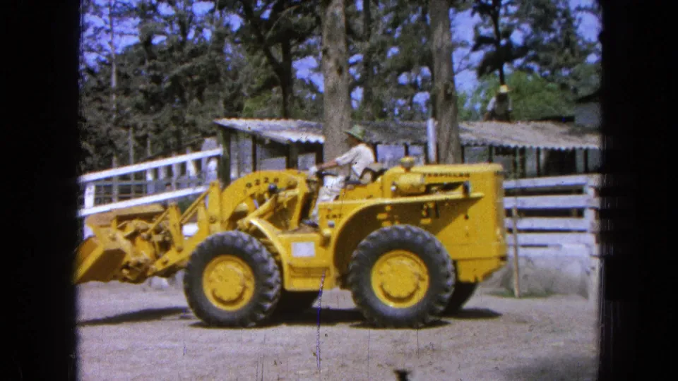 The 1965 8mm frame shows the loader closer to camera as the operator maneuvers it along the fenced dirt yard.