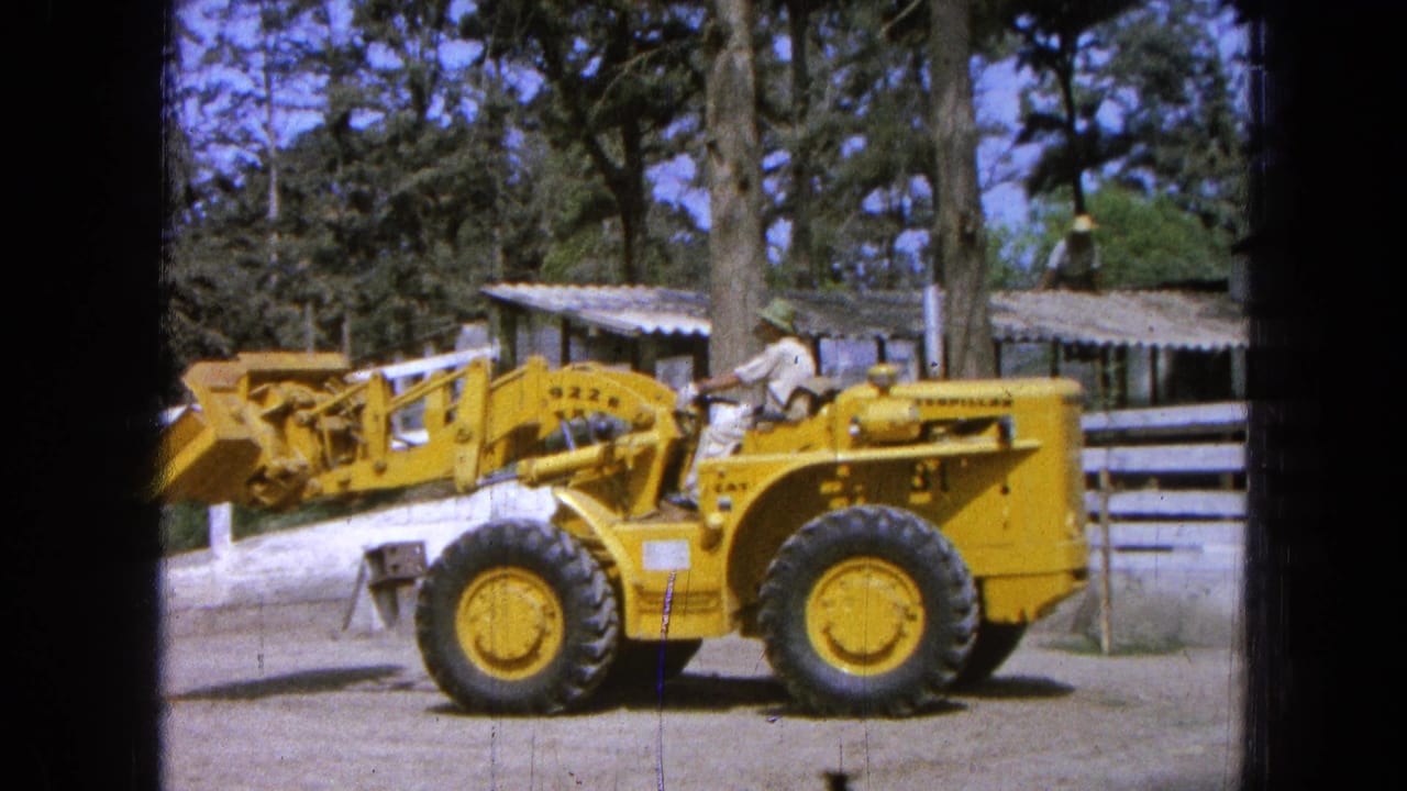 Yellow front-end loader beside a white fence with a seated operator and trees behind it.