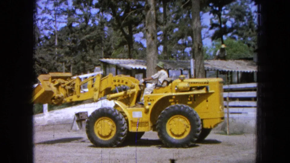 1965 Pueblo, Mexico home-movie frame of a yellow loader near a white fence, with the bucket low and an operator seated at the controls.