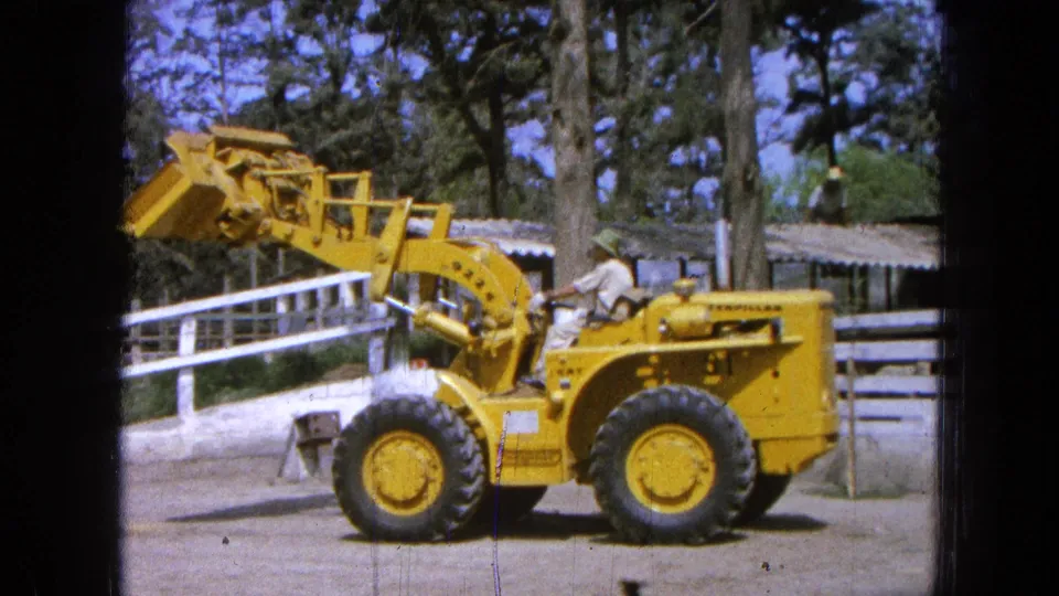 1965 Pueblo, Mexico home-movie frame of a yellow loader lifting its bucket higher, with the operator visible near a fenced rural site.