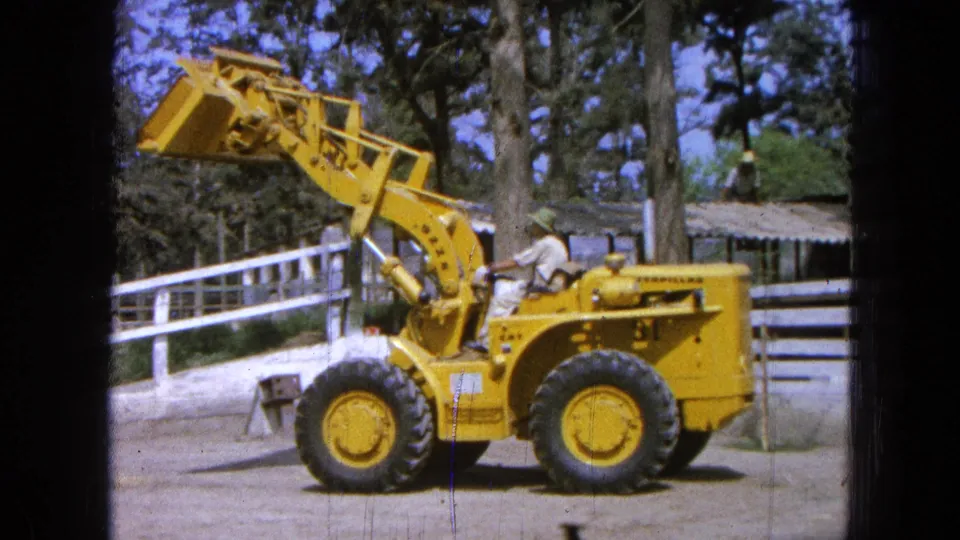 1965 Pueblo, Mexico frame showing a yellow Caterpillar loader with bucket held high, operator seated, and rural fencing behind.