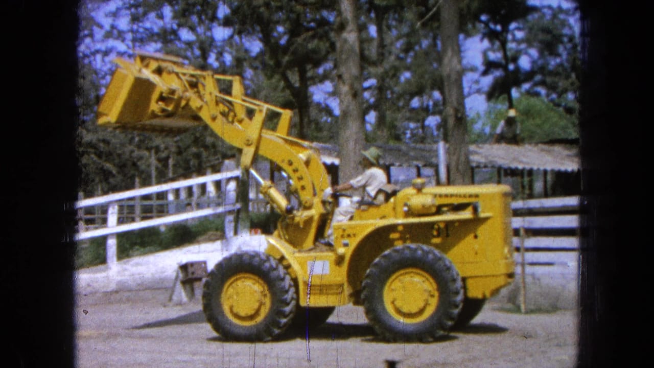 Yellow front loader with a raised bucket beside a white ranch fence and trees.