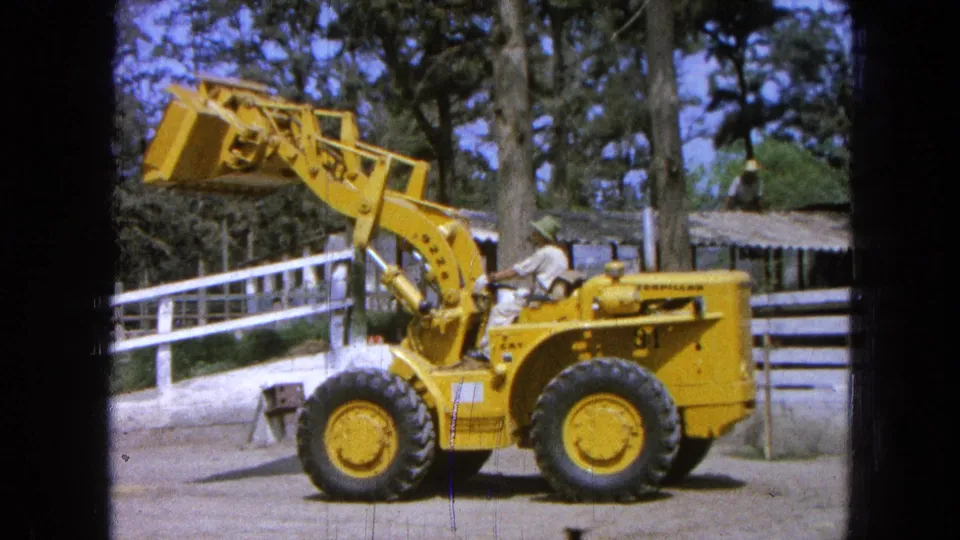 1965 Pueblo, Mexico 8mm frame showing a yellow loader driven past a white fence in a ranch yard.