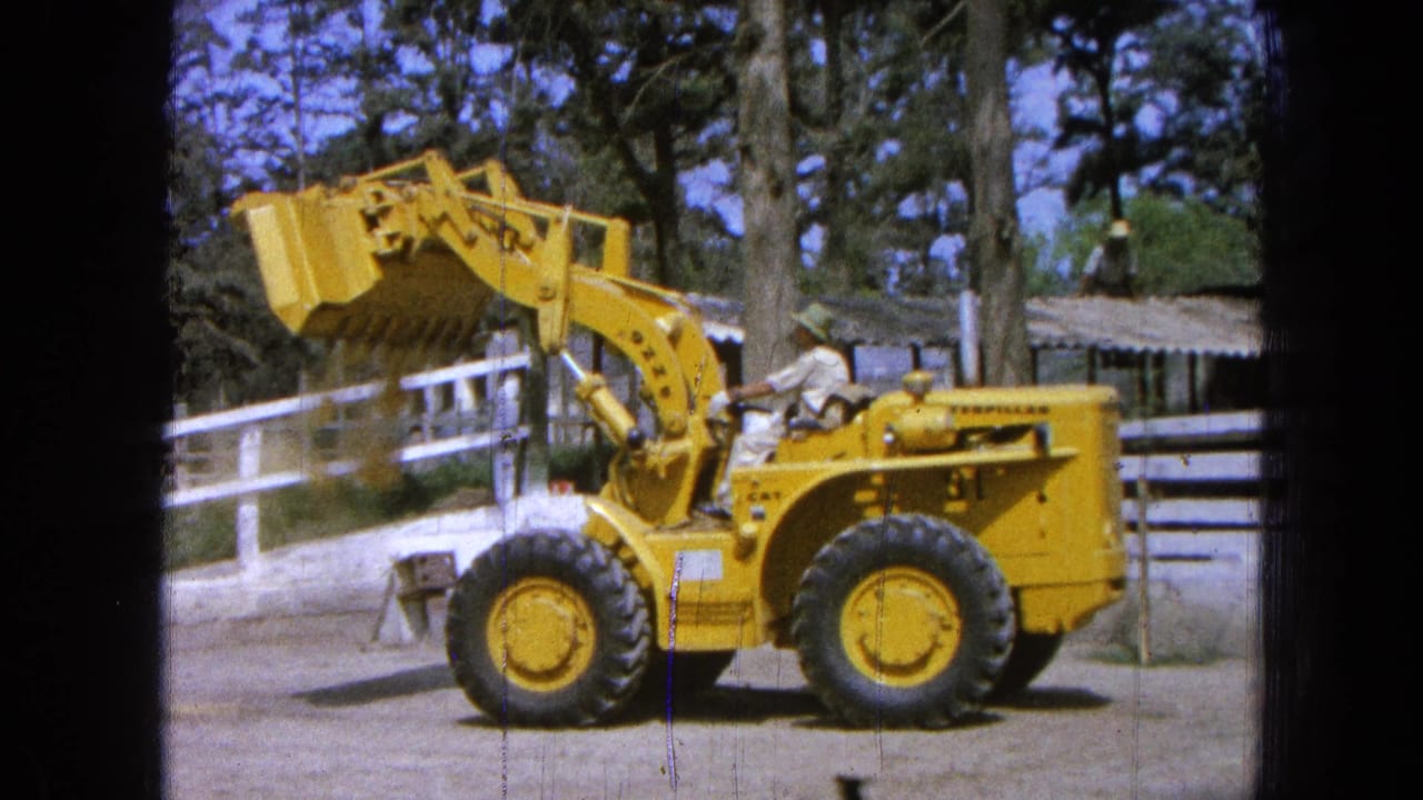 Yellow loader in profile with its bucket lifted near ranch fencing and trees.