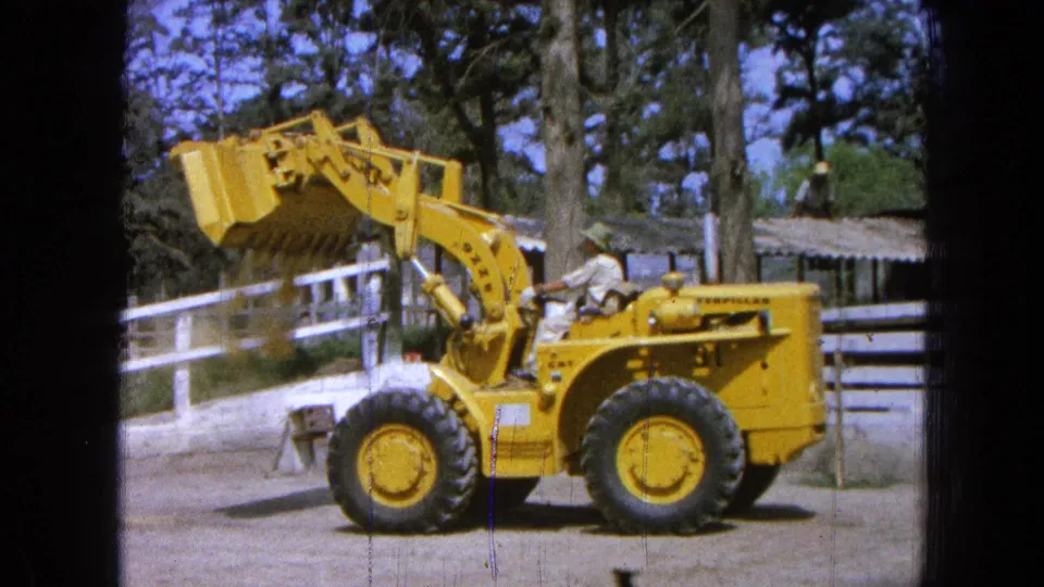 1965 Pueblo, Mexico frame of a yellow loader in profile, with the operator seated and the bucket raised beside ranch fencing.