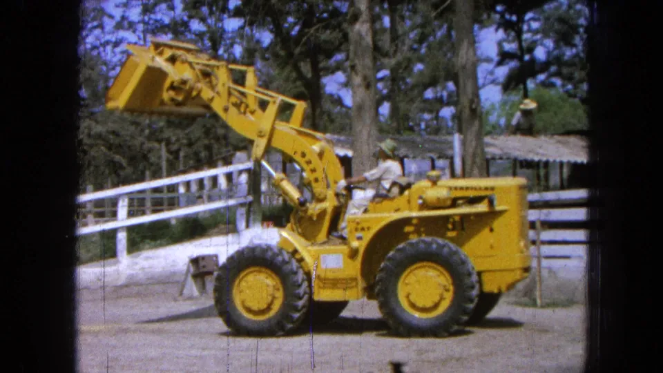 1965 Pueblo, Mexico 8mm frame showing the yellow loader and driver near a white fence and shaded ranch buildings.