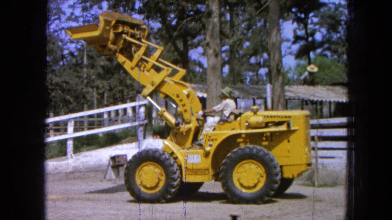 Yellow front-end loader with raised bucket beside a white fence and trees.