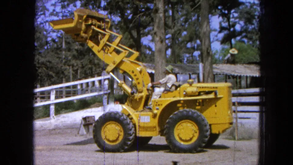 1965 Pueblo, Mexico 8mm frame showing a yellow front-end loader with its bucket raised and an operator seated near a white fence.