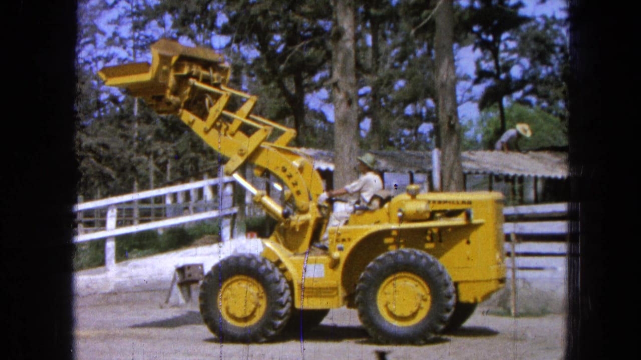 Operator on a yellow loader with raised bucket near a fence and shelter.