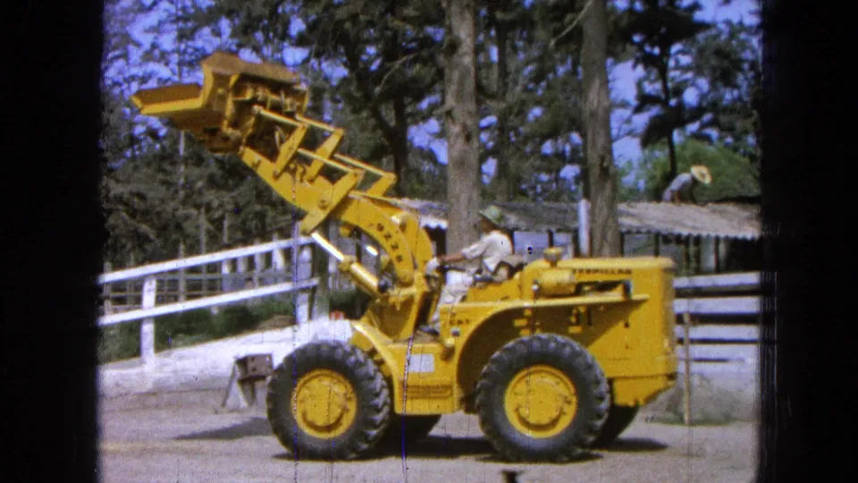 The yellow loader remains in profile in Pueblo, Mexico, with the operator seated and the bucket still held high near a white rail fence.