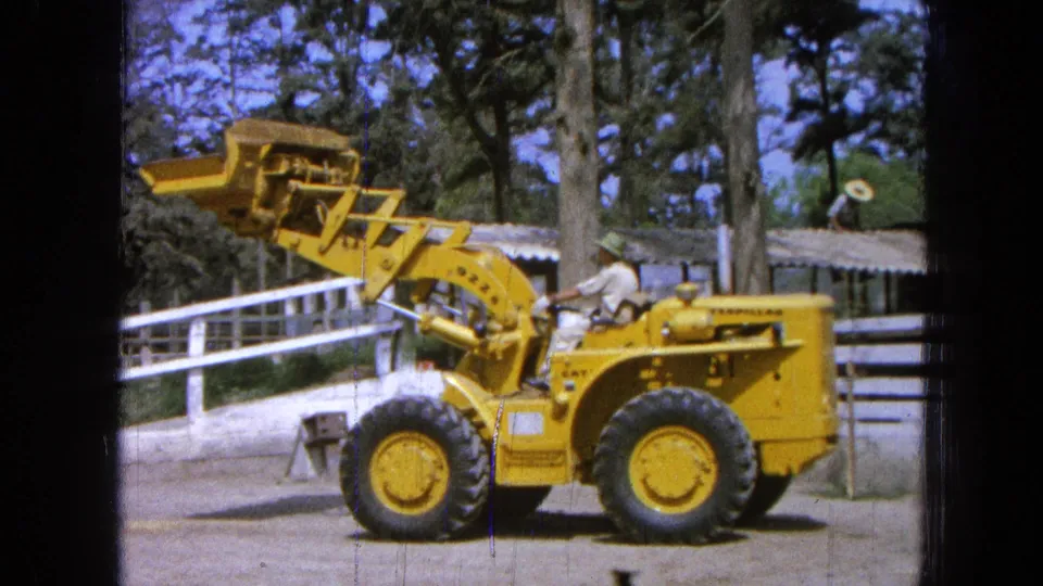 Mid-1960s 8mm footage shows the loader bucket partly lowered as the operator controls the yellow machine in a rural outdoor setting.