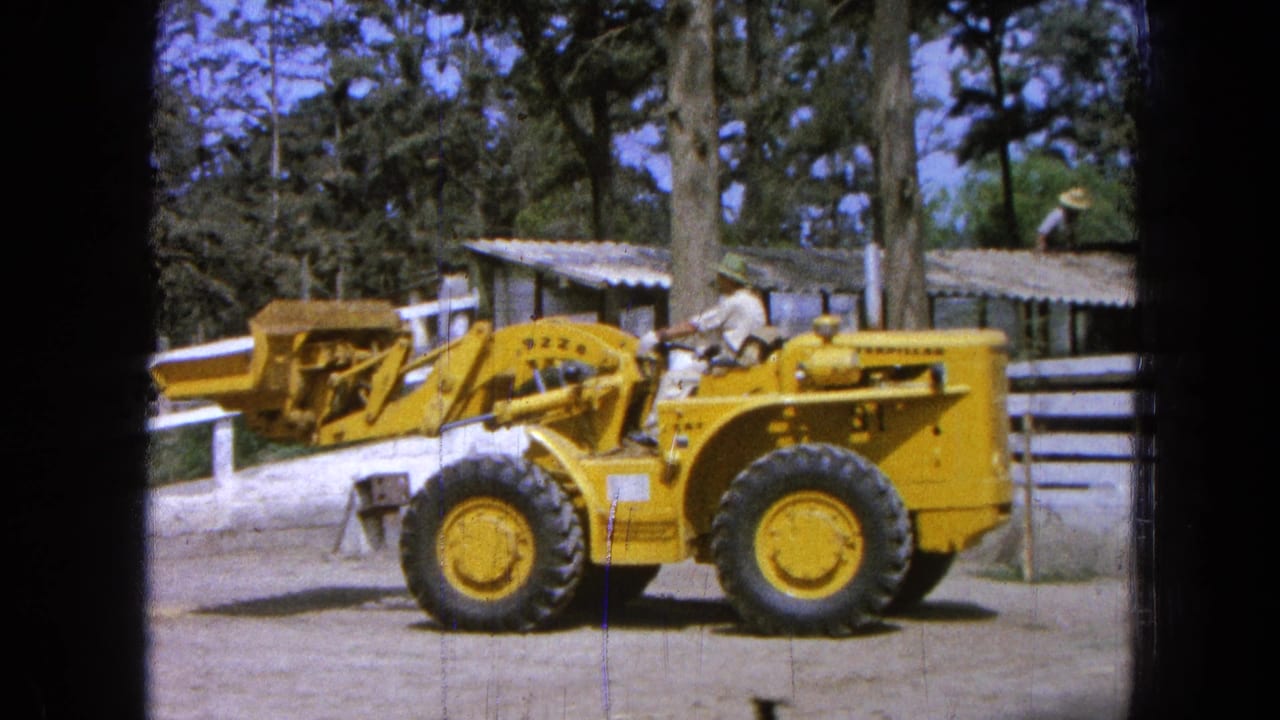 1965 Pueblo, Mexico 8mm frame showing a yellow front-end loader in profile near a white fence, trees, and low buildings.