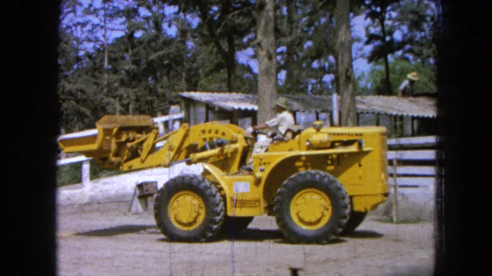 1965 Pueblo, Mexico 8mm frame showing a yellow front-end loader in profile near a white fence, trees, and low buildings.