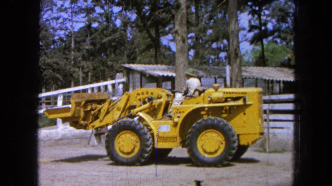1965 Pueblo, Mexico frame of a yellow loader beside a white fence, with an operator visible and the bucket extended forward.