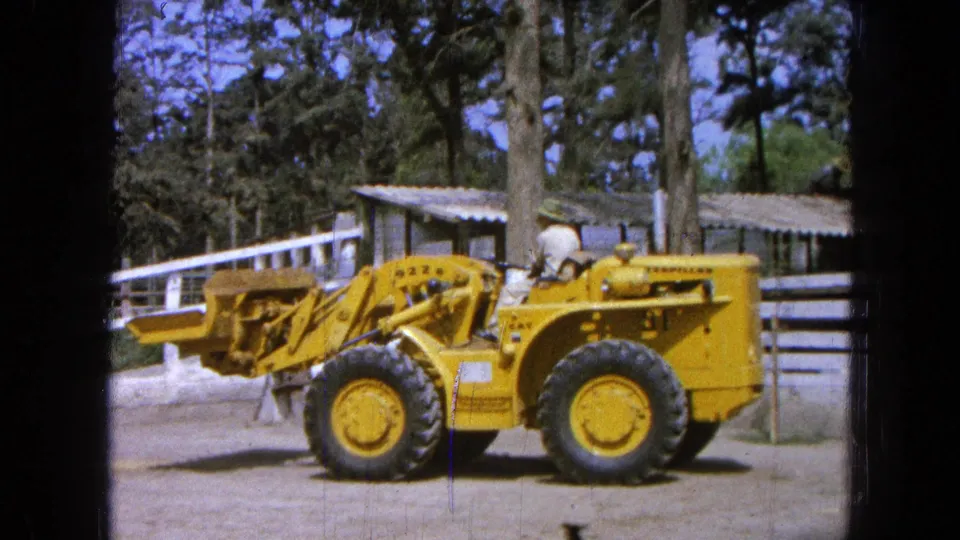 1965 Pueblo, Mexico frame of a yellow loader beside a white fence, with an operator visible and the bucket extended forward.