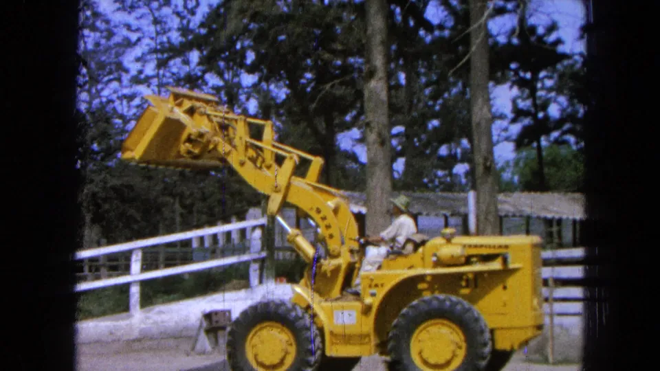 1965 Pueblo, Mexico frame showing the loader arm lifted and bucket raised, with the operator seated below and trees behind.