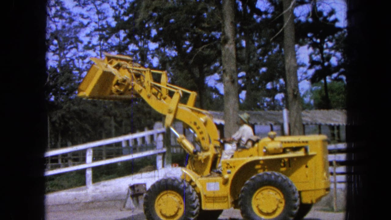 Yellow front-end loader with raised bucket beside a white fence and trees.