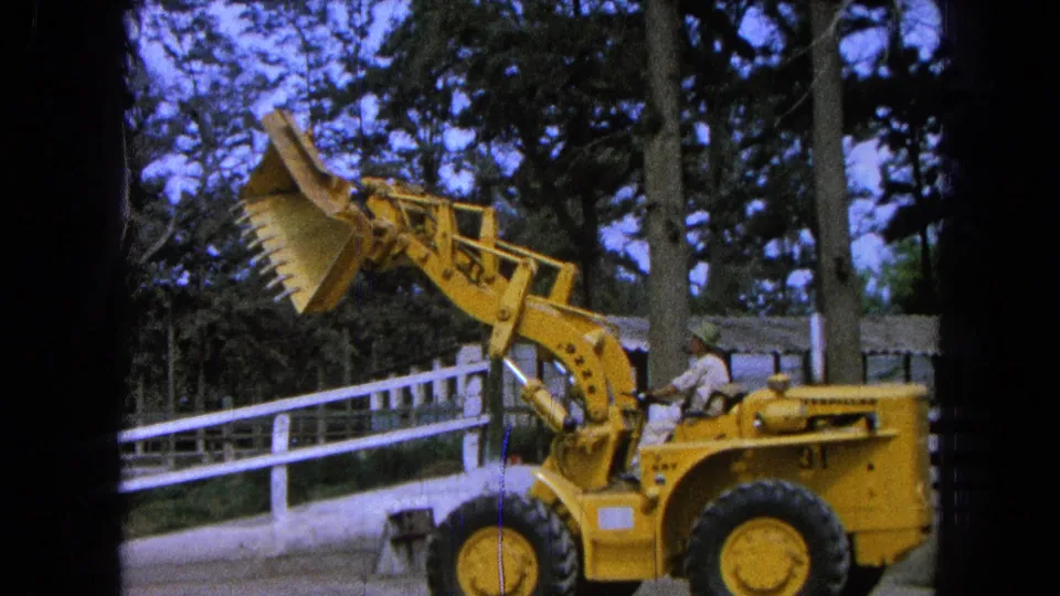A yellow front-end loader in 1965 Pueblo, Mexico is seen with the bucket tilted upward as the operator demonstrates its movement.