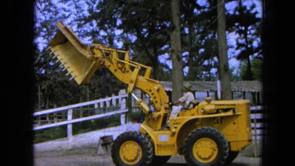 The operator raises the front-end loader bucket high near the white fence, with trees and film-frame edges visible.