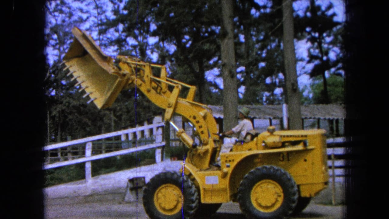 1965 Pueblo, Mexico home-movie frame showing a man seated on a yellow front-end loader with its bucket raised near a white fence and tall trees.