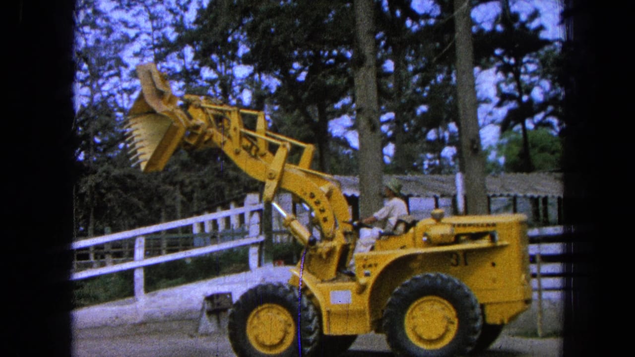 1965 Pueblo, Mexico frame of a yellow loader seen from the side, with the operator visible and the bucket raised in front of a white fence.