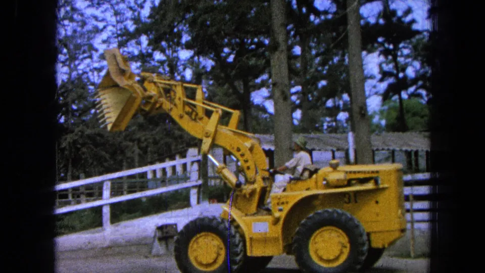 1965 Pueblo, Mexico frame of a yellow loader seen from the side, with the operator visible and the bucket raised in front of a white fence.
