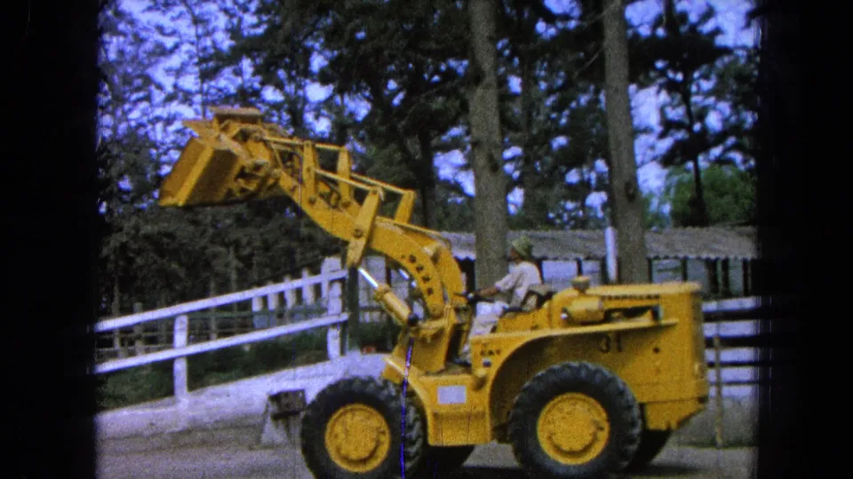 1965 Pueblo, Mexico frame showing the yellow front-end loader near the end of the clip, with operator, raised bucket, fence, and wooded background.