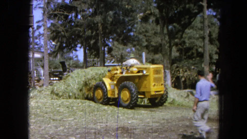 A yellow loader works near a large hay pile while an adult stands at the edge of the rural yard in the 1965 Pueblo, Mexico footage.