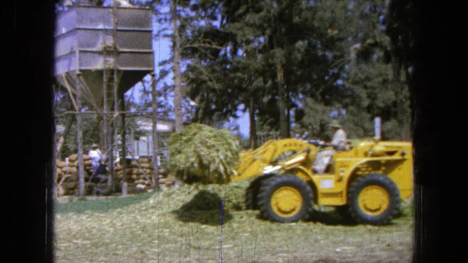 A yellow front-end loader moves across the yard with green grass lifted, with a silo-like structure and trees in the background.