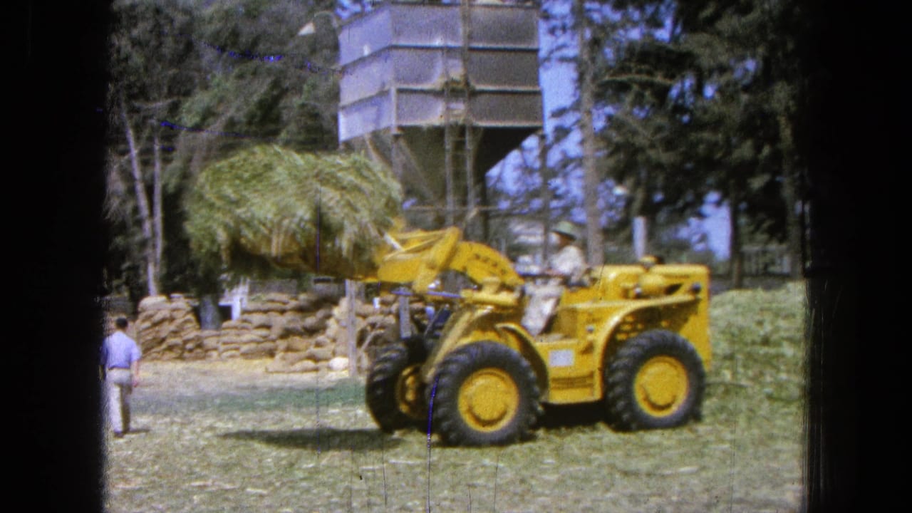 Yellow loader lifts a bundle of hay near stacked bales and a metal silo.