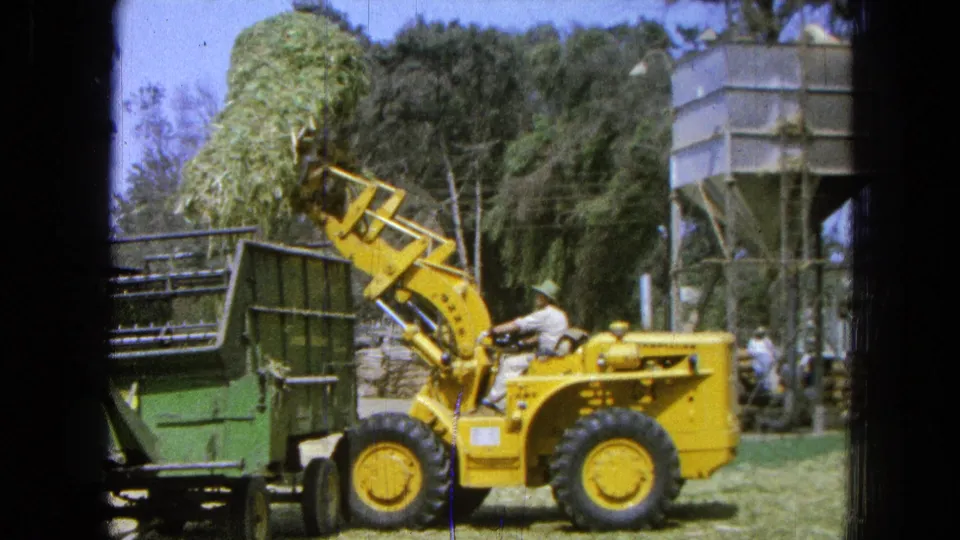 A farm worker operates the yellow loader with hay lifted over a green trailer in the 1965 Pueblo, Mexico farm scene.