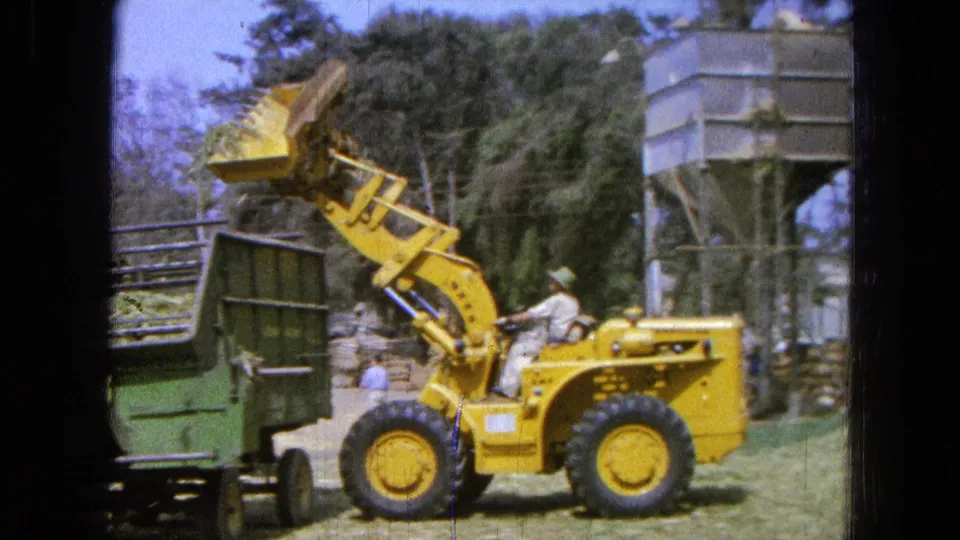 A yellow loader carries dry grass above a green trailer in Pueblo, Mexico, with the seated operator and background silo visible.