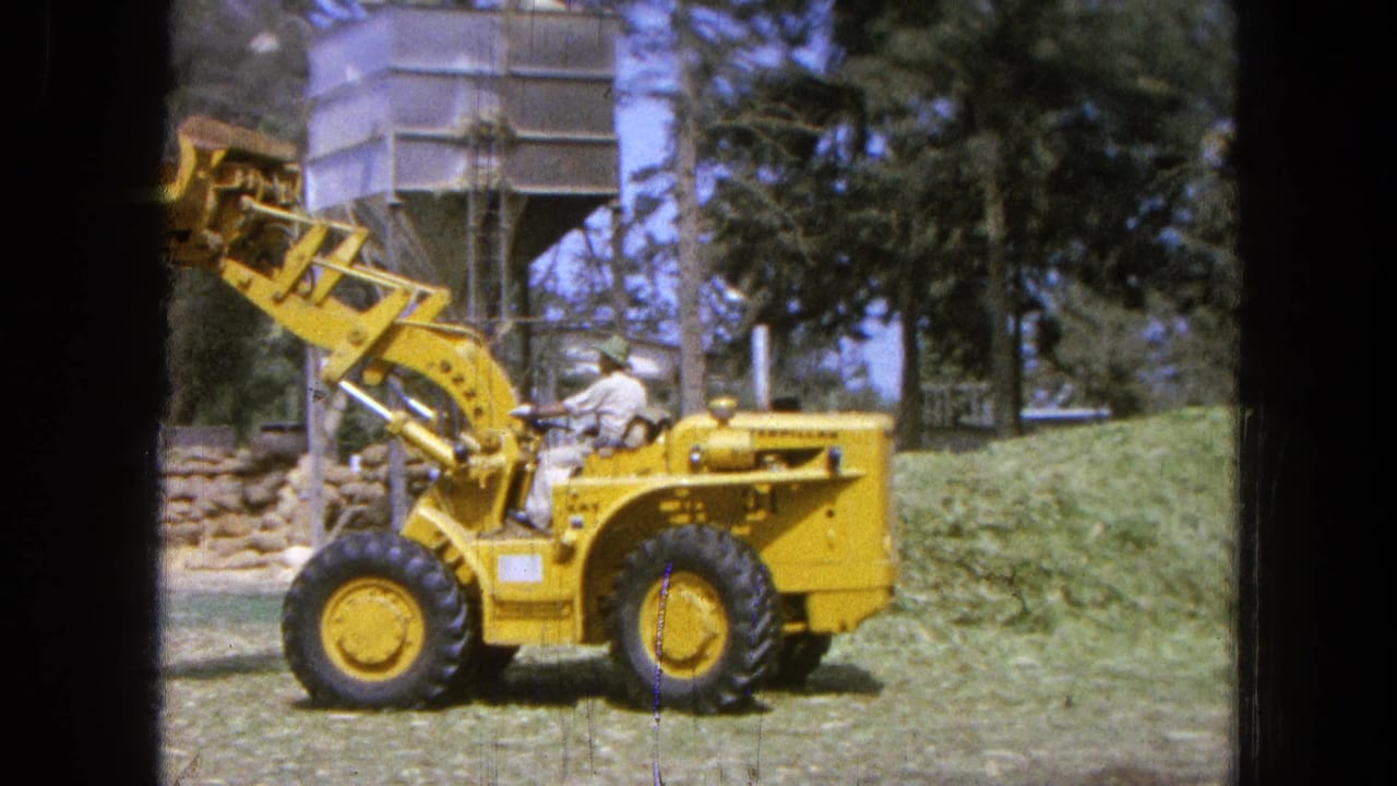 Yellow front-end loader with raised bucket beside cut grass and trees.