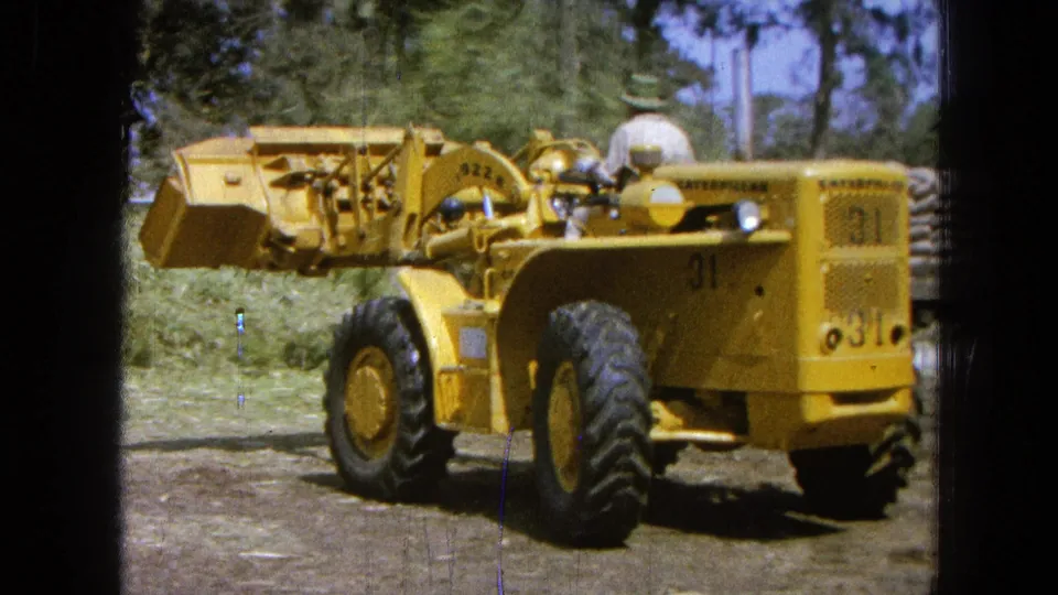The loader is framed from behind as the bucket sits raised with cut grass nearby, showing the operator during outdoor machinery work.