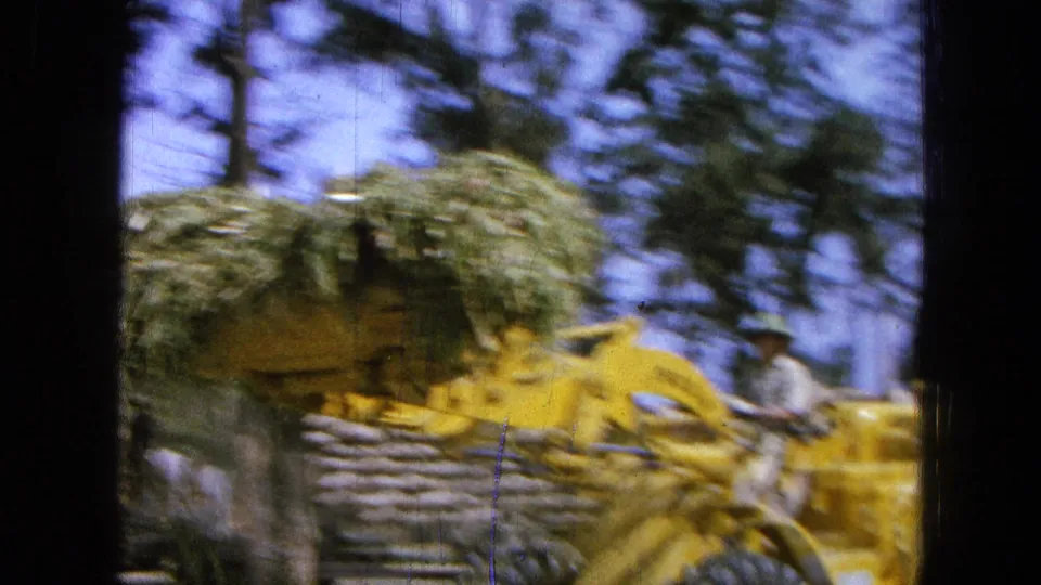 Motion-blurred 8mm frame of the yellow loader moving with a load of green hay or straw, with trees behind it.