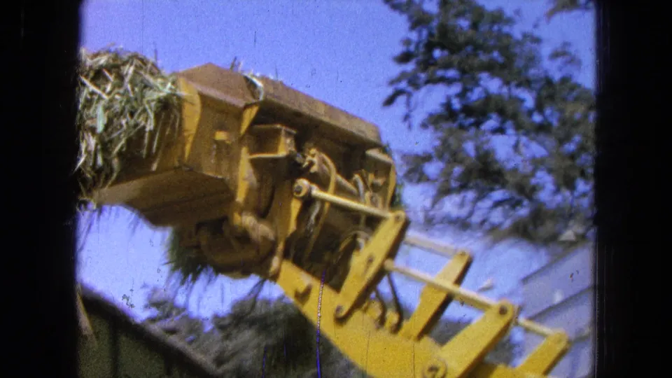 A yellow agricultural loader tilts a hay-filled bucket, with trees and sky visible in the 1965 Pueblo, Mexico scene.