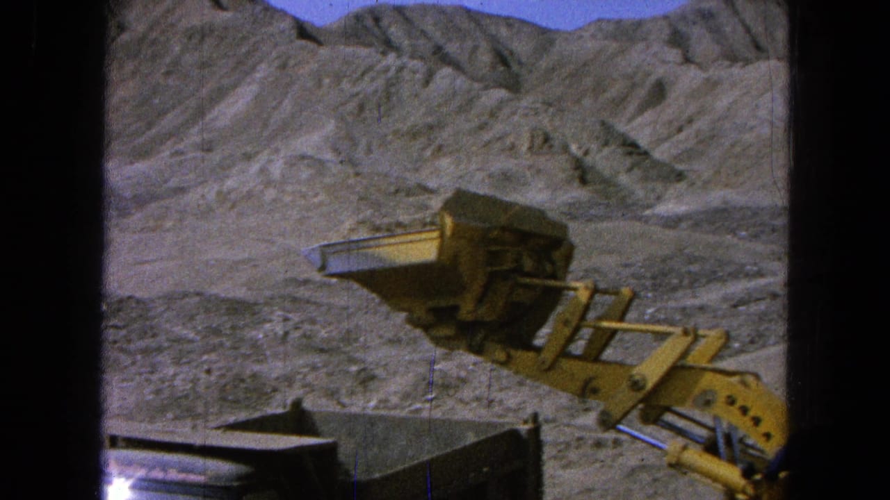 Loader bucket positioned above a dump truck bed in a dry rocky landscape.